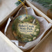 A circular ornament with a painting of a forest and a waterfall, placed in a box with a white tissue paper lining.
