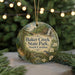 A round ornament with a painting of a bird and a waterfall, surrounded by greenery, hangs in front of a Christmas tree with lights.