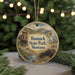 A round ornament with a painting of a rural landscape, featuring a building, a wagon, and a stream, hanging from a string.