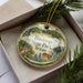A decorative ornament in a box, featuring a scenic image of Bastrop State Park, Texas, with a deer and other wildlife elements.