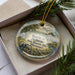 A decorative ornament in a box, featuring a scene depicting a historical battle site with a cannon and a wooden fence.