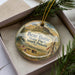 A decorative ornament in a box, featuring a scenic landscape with a mountain and pine trees, along with the text "Bear Butte State Park, South Dakota".