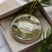 A decorative ornament with a scenic image of Beartown State Park West Virginia, featuring a river, mountains, and a cabin, is displayed in a box with pine branches.