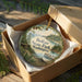 A decorative ornament in a box, featuring a scenic landscape of Belfair State Park in Washington.