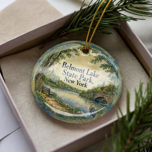 A round ornament with a scenic landscape of Belmont Lake State Park, New York, displayed in a box with pine branches.