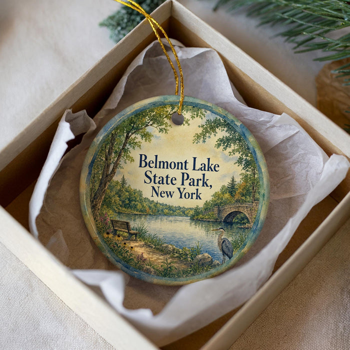 A round ornament with a painting of Belmont Lake State Park, New York, displayed in a box with a white tissue paper lining.