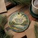 A round ornament with a painting of Belmont Lake State Park, New York, hangs on a wooden surface, surrounded by pine branches and a cup of coffee.