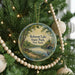 A round ornament hanging on a Christmas tree, featuring a scenic landscape with a lake, trees, and a bridge.
