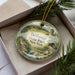 A decorative ornament in a box, featuring a scenic landscape with a bridge, trees, and a bird, with the text "Bethpage State Park, New York" prominently displayed.