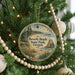 A round ornament hanging on a Christmas tree, featuring a scenic landscape with a beach, mountains, and a body of water.