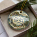 A decorative ornament in a box, featuring a scenic landscape with a lake, mountains, and pine trees.