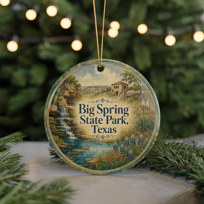 A round ornament with a painting of a scenic landscape, featuring a waterfall and a house, is displayed in front of a blurred background with Christmas lights.