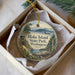 A decorative ornament in a white box, featuring a scenic landscape with mountains, a lake, and a wooden cabin.
