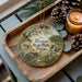 A decorative ornament featuring a scenic landscape of Blanco State Park, Texas, with a house, trees, and a waterfall. The ornament is displayed on a wooden tray, accompanied by pine cones and a lit candle.
