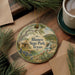 A round ornament with a scenic illustration of Blanco State Park, Texas, featuring a house, trees, and a river, is displayed on a wooden surface alongside a cup of coffee and some pine branches.