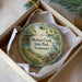A round ornament with a scenic image of a park, featuring a bird, a path, and a body of water, is displayed in a box.