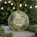A round ornament with a painting of a bird and a path, surrounded by greenery, hangs in front of a blurred background with twinkling lights.