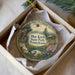 A decorative ornament in a box, featuring a scenic landscape with a bird perched on a branch, surrounded by flowers and greenery.
