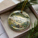 A decorative ornament in a box, featuring a scenic landscape with a house, trees, and mountains.