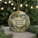 A circular ornament with a scenic image of a train station and a waterfall, surrounded by greenery, hangs in front of a Christmas tree with twinkling lights.