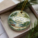 A decorative ornament in a box, featuring a scenic landscape with a lake, trees, and a boat, with the text "Bomosoen State Park, Vermont" printed on it.