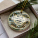 A decorative ornament in a box, featuring a scenic landscape with mountains, waterfalls, and greenery.