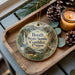 A wooden tray with a decorative ornament featuring a scenic image of the Booth State Scenic Corridor, Oregon, along with pine cones and a lit candle.