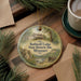 A round ornament with a painting of a lodge and a river, surrounded by pine branches and a cup of coffee on a wooden surface.