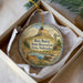 A round ornament with a scenic landscape featuring a mountain, windmill, and deer, placed in a white box with a gold string.
