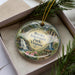 A decorative ornament in a box, featuring a scenic landscape with a waterfall and pine trees.