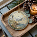 A decorative ornament featuring a lighthouse and coastal scenery, placed on a wooden tray alongside pine cones and a lit candle.