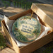 A round ornament in a wooden box, featuring a scenic image of a forest and a path, with the text "Bridle Trails State Park Washington" printed on it.