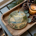 A wooden tray with a decorative ornament featuring a scenic beach landscape, pine cones, and a lit candle.