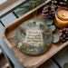 A decorative ornament featuring a scenic landscape of Brooks Memorial State Park in Washington, with pine cones, a candle, and greenery surrounding it.