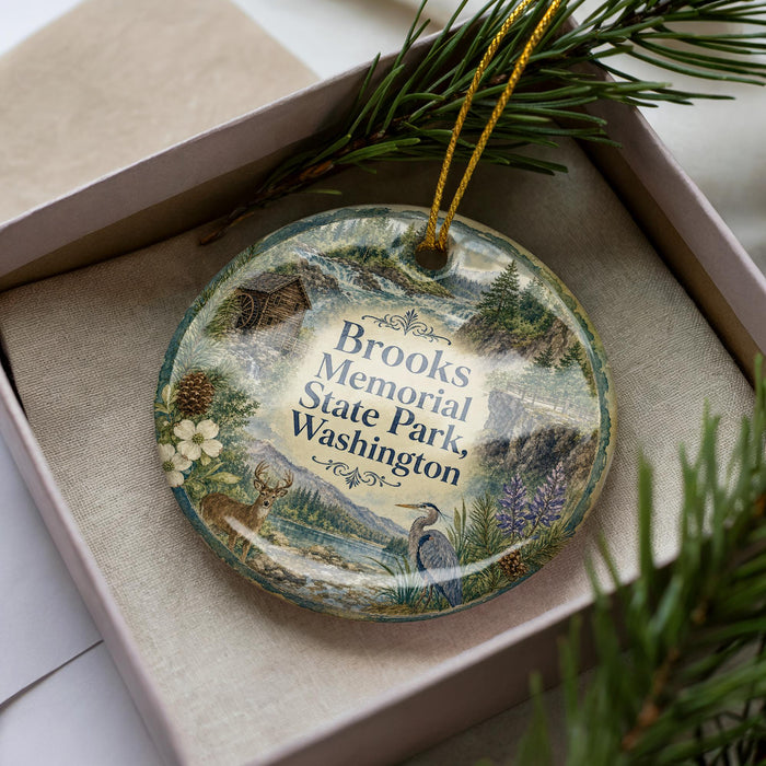 A decorative ornament in a box, featuring a scenic landscape with mountains, pine trees, and wildlife.