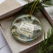 A decorative ornament in a box, featuring a scenic landscape with mountains, pine trees, and wildlife.