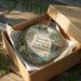 A decorative ornament in a wooden box, featuring a scenic landscape with a river, trees, and wildlife.