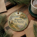 A round ornament with a scenic landscape and text, hanging on a wooden surface next to a cup of coffee and pine branches.