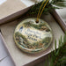 A decorative ornament in a box, featuring a scenic landscape with a house, trees, and a deer.