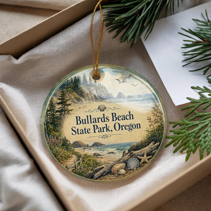 A decorative ornament with a scenic beach landscape, featuring a path, trees, and a body of water, is displayed in a box with pine branches.