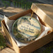 A decorative ornament in a wooden box, featuring a scenic landscape with a beach, mountains, and wildlife.