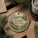 A round ornament with a scenic landscape of burlingame state park, Rhode island, is displayed on a wooden surface alongside a cup of coffee and some pine branches.