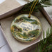 A decorative ornament in a box, featuring a scenic landscape with a cabin and trees, with the text "Burr Oak State Park, Ohio" printed on it.