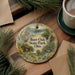A decorative ornament with a scenic landscape of Burr Oak State Park, Ohio, featuring a cabin and a waterfall, is displayed on a wooden surface alongside a cup of coffee and some pine branches.