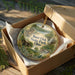 A decorative ornament in a brown box, featuring a scenic landscape with a cabin and trees, with the text "Burr Oak State Park, Ohio" printed on it.