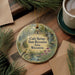 A decorative ornament featuring a scenic landscape with a bird, surrounded by pine branches and a cup of coffee on a wooden surface.