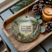 A decorative ornament featuring a scenic landscape of Caledonia State Park, Pennsylvania, with a waterfall and pine trees. The ornament is placed on a wooden tray, accompanied by a lit candle and pine cones.