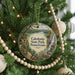 A round ornament hanging on a Christmas tree, featuring a scenic landscape with a waterfall and a path leading to a cabin.