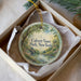 A decorative ornament in a box, featuring a scenic landscape with a river, trees, and a bridge.