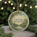 A round ornament with a painting of a park scene hangs in front of a Christmas tree with lights.